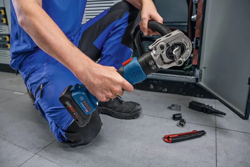 A person wearing safety equipment uses a cordless hydraulic crimping tool near electrical cabinets.