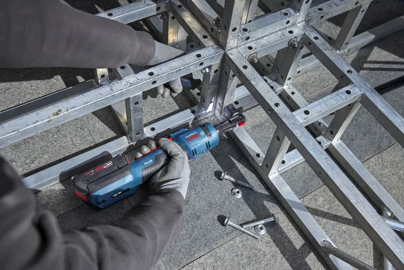Person wearing safety equipment tightens bolts on a metal frame using a cordless ratchet wrench.