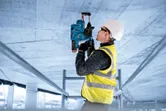 A person wearing safety equipment operates a rotary hammer drill on a concrete ceiling.