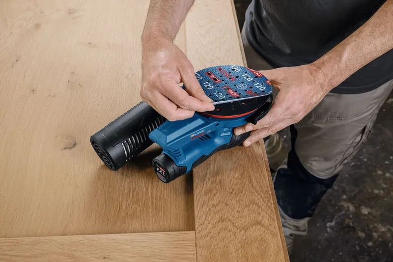 A person adjusts a sanding pad on a cordless random orbit sander near a wooden surface.
