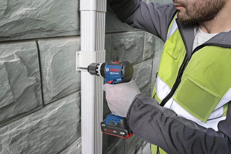 A person wearing safety equipment uses a cordless drill to fasten a bracket on a wall.