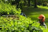A person wearing safety equipment trims a hedge with a cordless hedgecutter.