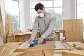 A person wearing safety equipment sands a wooden panel with a multifunction tool.