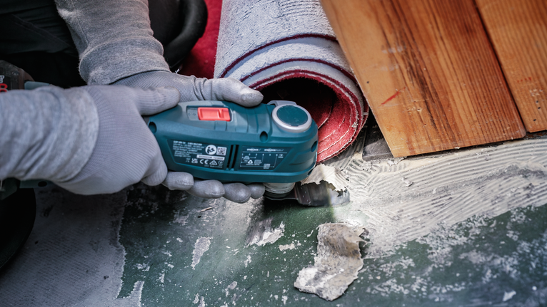 Person wearing safety equipment uses a power tool to remove adhesive near rolled carpet.