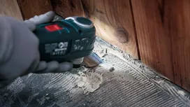 Person wearing safety equipment scrapes adhesive from a floor near wooden wall.