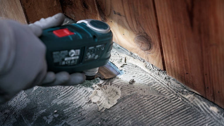 Person wearing safety equipment scrapes adhesive from a floor near wooden wall.