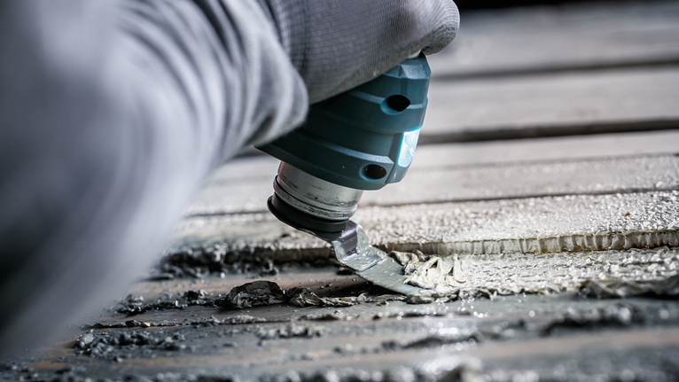 A person wearing safety equipment uses a power tool to remove adhesive from a surface.