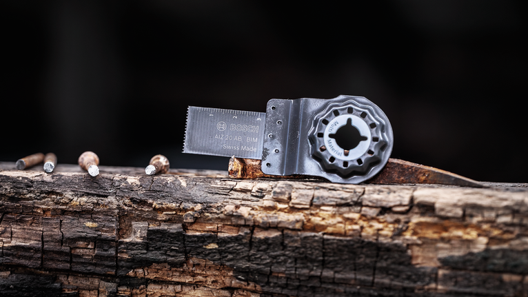 Oscillating multi-tool blade resting on a weathered wooden beam beside four nails.