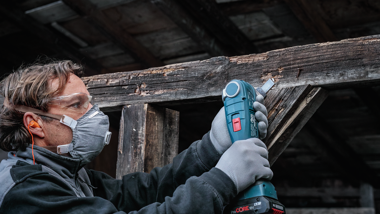 Person wearing safety equipment uses a cordless tool to sand rough wood.