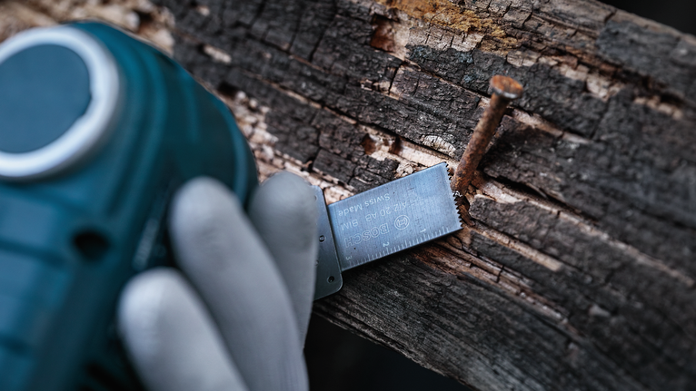 A person wearing safety equipment cuts through wood and a nail with a power tool.