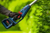 A person wearing safety equipment trims hedges with a cordless hedgecutter.
