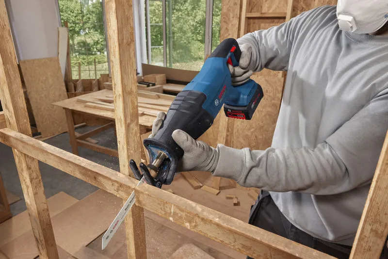 A person wearing safety equipment cuts wood framing with a cordless reciprocating saw.