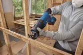 A person wearing safety equipment cuts wood framing with a cordless reciprocating saw.