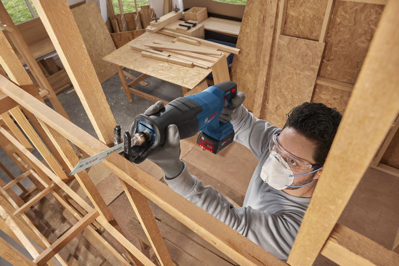 A person wearing safety equipment uses a cordless reciprocating saw to cut wood.