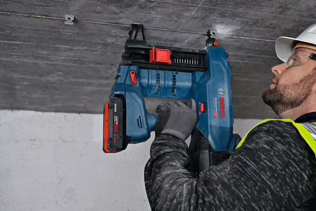 A person wearing safety equipment drills into a ceiling to install a metal hanger.
