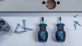 Two screwdrivers and several screws arranged on a white surface near a drilled board.