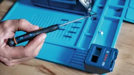 Person uses a screwdriver to hold a screw near a blue storage case on a wooden surface.