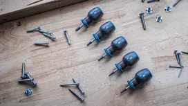 Six blue and black screwdrivers on a wooden surface beside scattered screws and nuts.