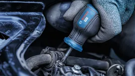 Person wearing safety equipment tightens a screw on a car engine with a screwdriver.