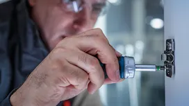 Person wearing safety equipment using a power screwdriver to fasten a hinge on a cabinet.