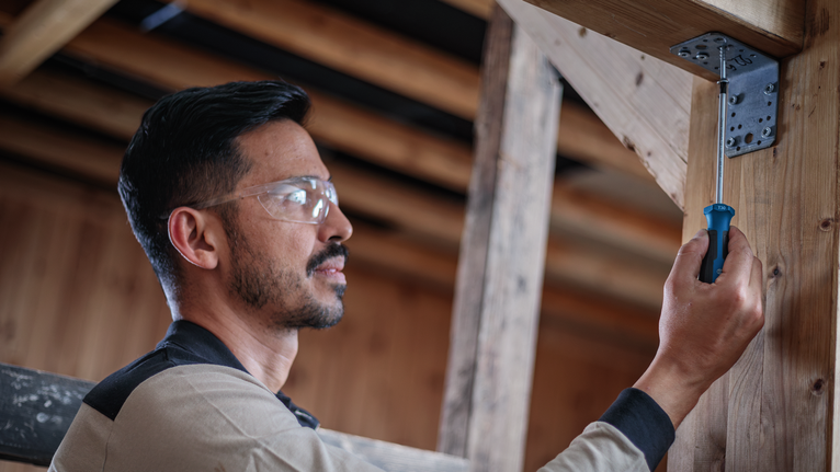 Person wearing safety equipment tightens a screw in a metal bracket on a wooden beam.