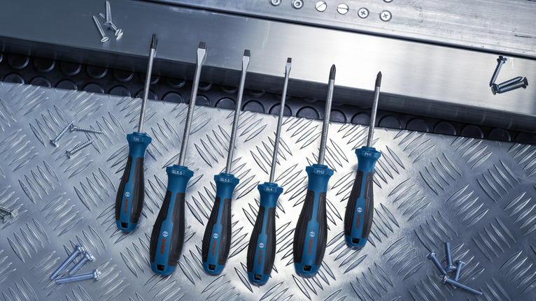 Six screwdrivers with blue handles arranged on a metal surface near scattered screws.