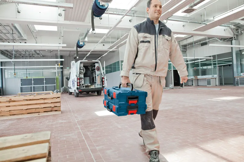 A person wearing safety equipment carries stacked toolboxes in a spacious workshop.