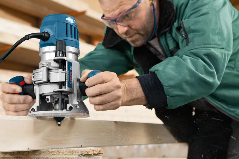 A person wearing safety equipment shapes wood with a handheld router on a workbench.