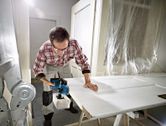 A person wearing safety equipment uses a cordless planer to smooth a white door.