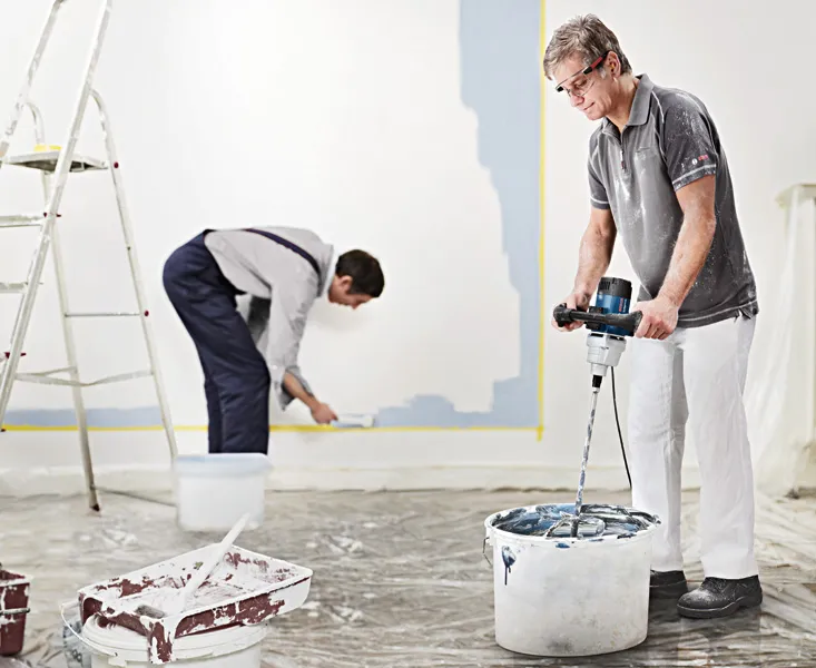 A person wearing safety equipment mixes paint with a stirring mechanism, while another paints a wall.