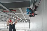 A person wearing safety equipment aligns ceiling frames using a laser leveling tool on a universal mount.