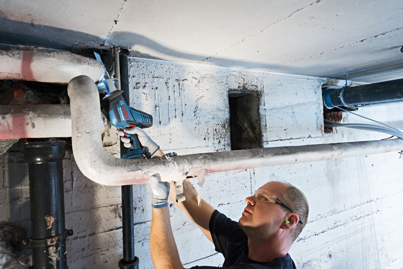 A person wearing safety equipment cuts a ceiling pipe with a power tool.