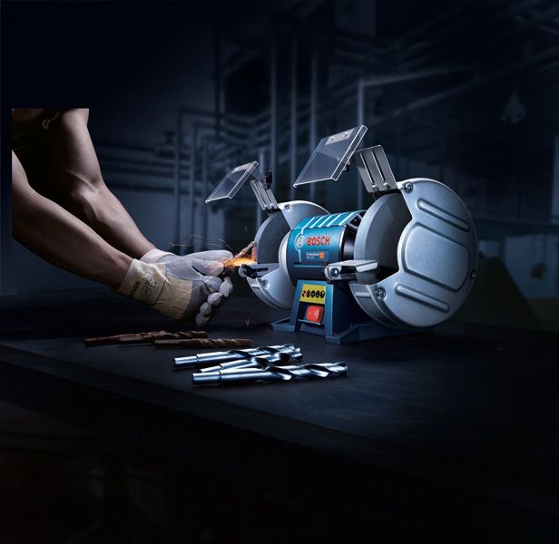 A person wearing safety equipment sharpens a drill bit with a double-wheeled bench grinder.