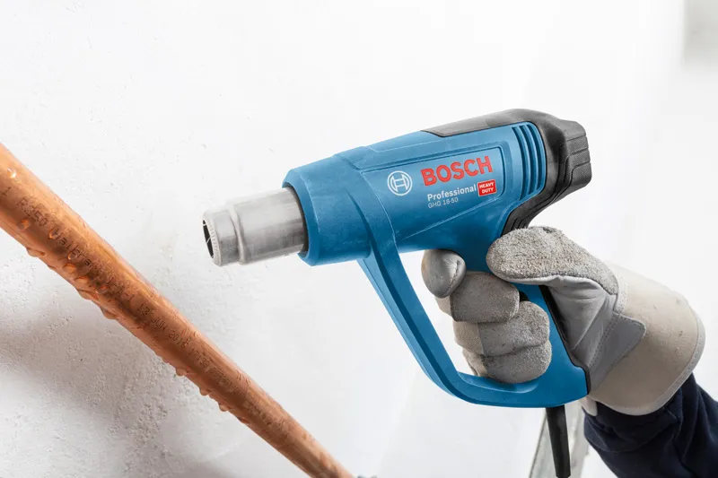 A person wearing safety equipment uses a blue heat gun near a copper pipe.