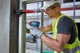 A person wearing safety equipment uses a cordless drill to fasten a window frame.