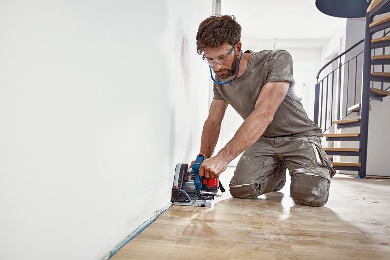 Person wearing safety equipment cuts along the floor near a wall with a power saw.