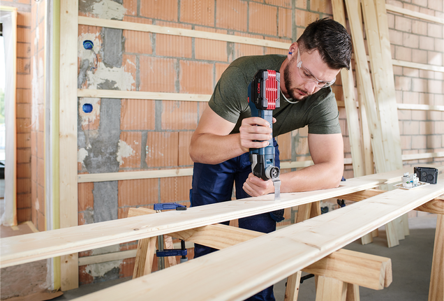 Person wearing safety equipment uses a power tool to cut a wooden plank on a workbench.