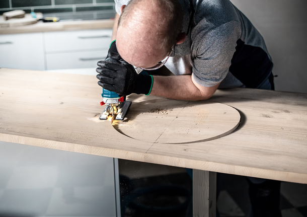 Person wearing safety equipment cuts a circular shape into wood with a jigsaw.