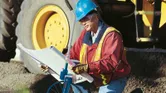 A person wearing safety equipment uses a measuring wheel while inspecting blueprints.