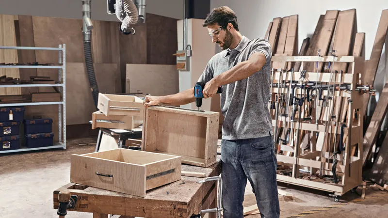 A person wearing safety equipment uses a cordless drill on a wooden drawer in a workshop.