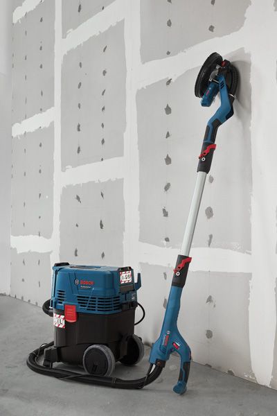 Drywall sander connected to a dust extractor standing against an unfinished wall.
