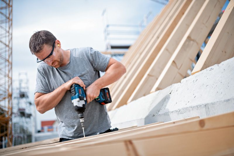 Person wearing safety equipment uses a cordless impact wrench to drive a screw into wood.