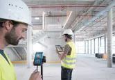 Two people wearing safety equipment use a laser measure and tablet in a building under construction.