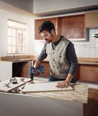 Person wearing safety equipment drills into a wooden board in a kitchen workspace.