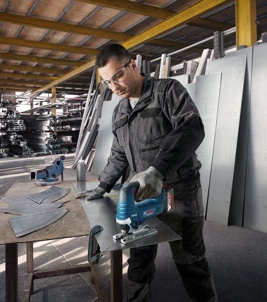 A person wearing safety equipment uses a cordless jigsaw to cut sheet metal in a workshop.