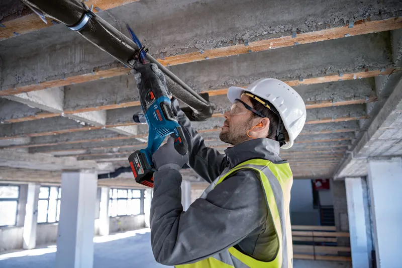 A person wearing safety equipment uses a reciprocating saw to cut a ceiling pipe.