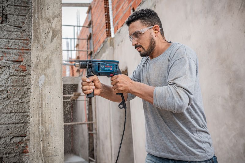 A person wearing safety equipment drills into a concrete wall with an impact drill.