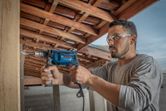 A person wearing safety equipment drills into a wooden beam with an impact drill.