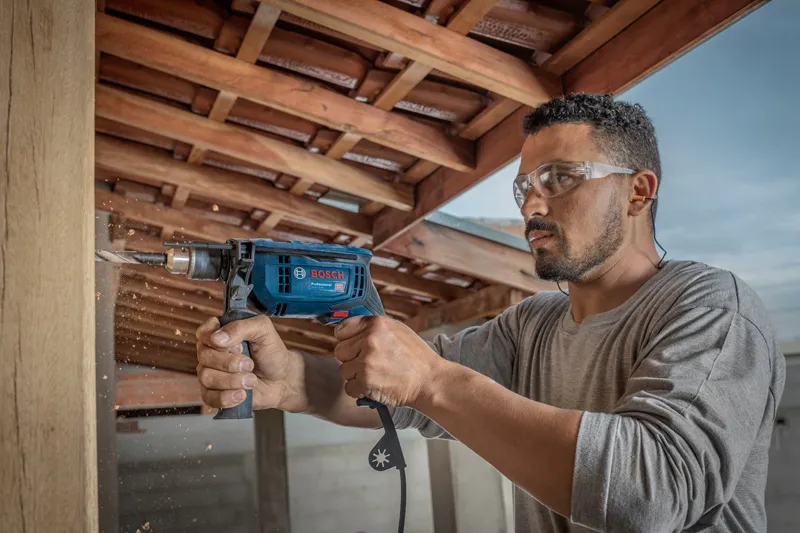 A person wearing safety equipment drills into wood under a wooden roof.