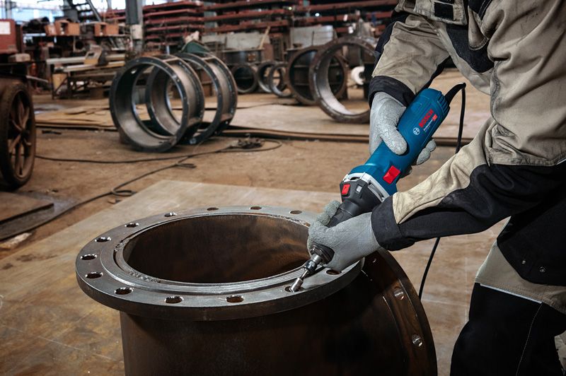 A person wearing safety equipment grinds the edge of a large metal pipe with a straight grinder.